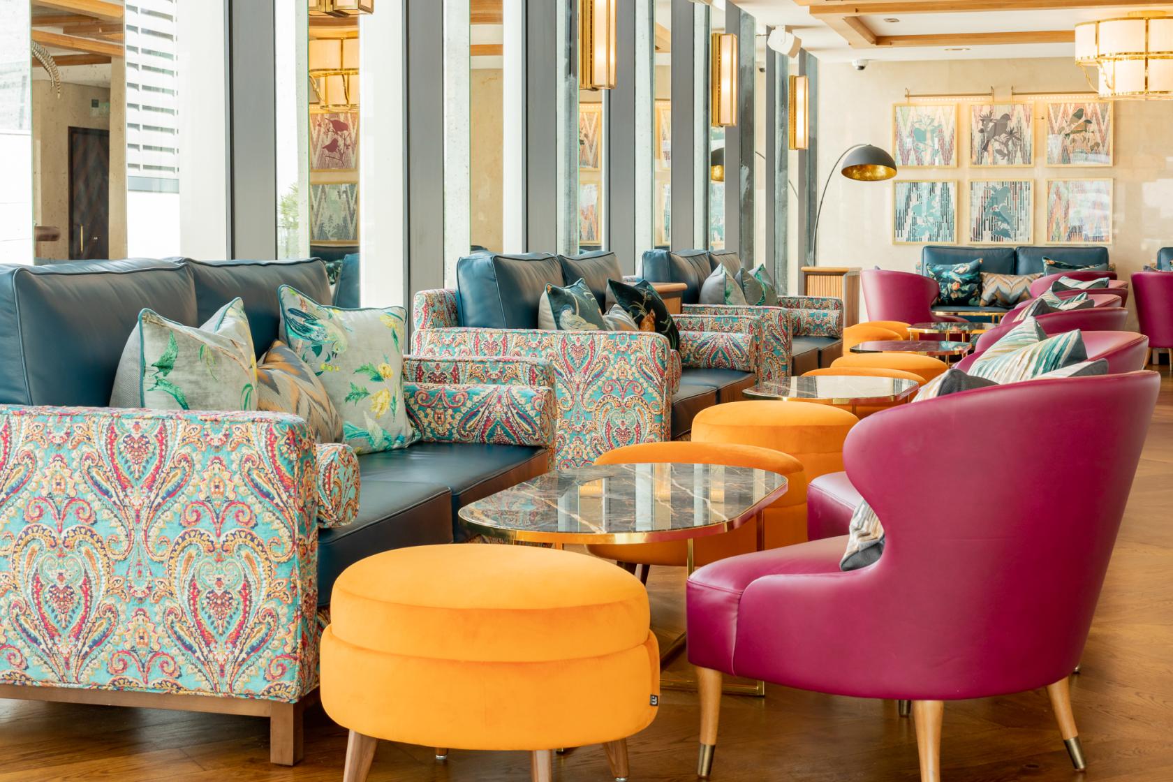A lounge with patterned armchairs, colorful stools, and large windows at Aviary Rooftop Bar & Restaurant.