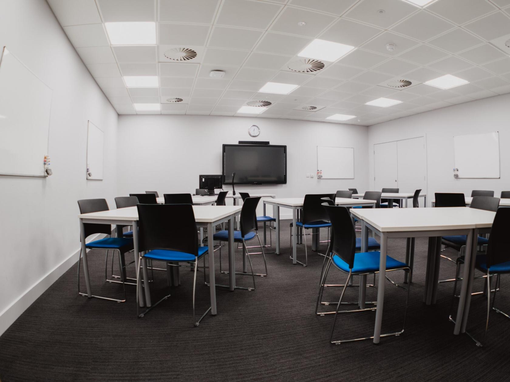 A meeting room with modular tables, blue and black chairs, wall-mounted whiteboards, and a large screen at University of Law - Leeds.