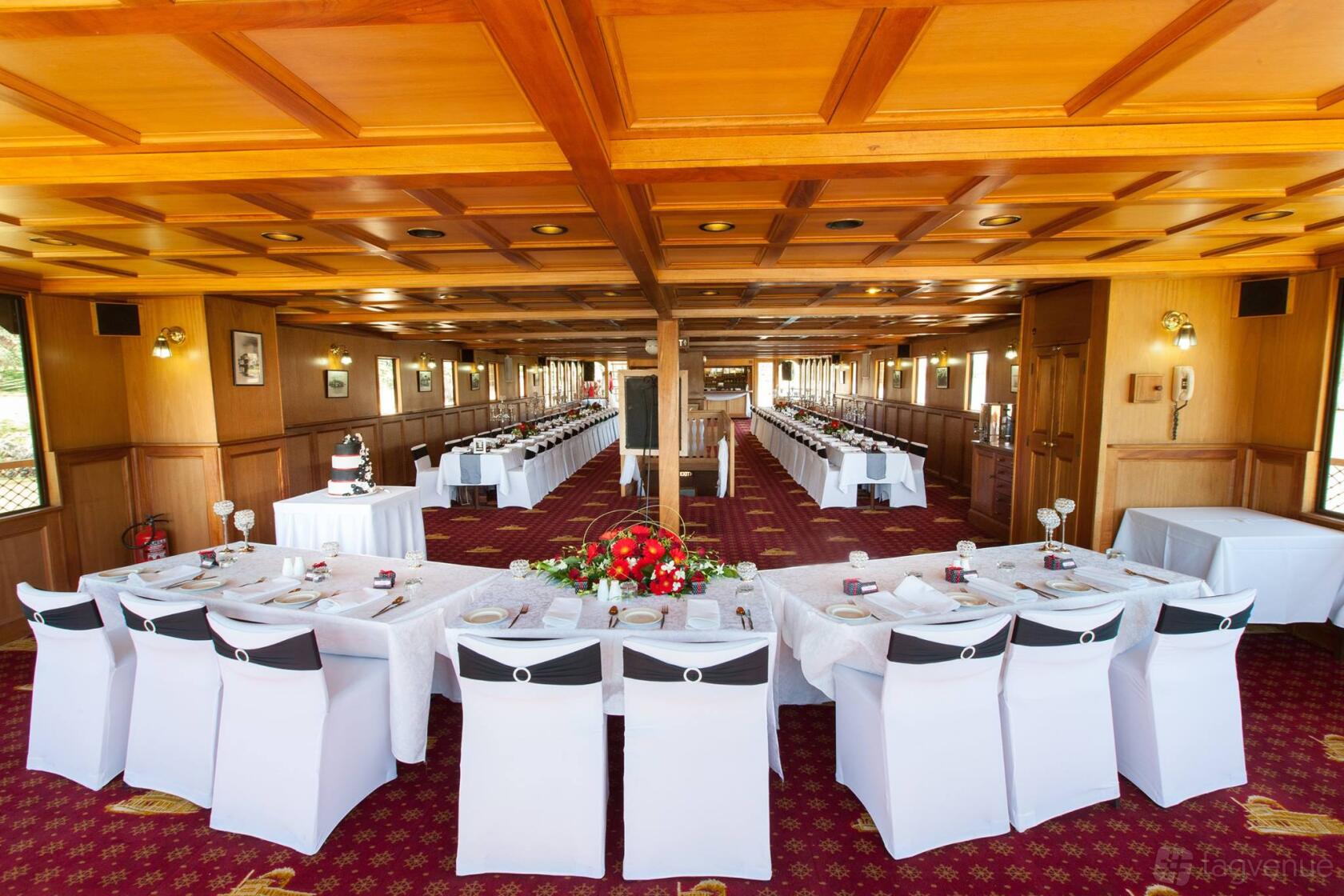 An event venue with wood-paneled ceilings, large windows, and long tables with white linens at Mildura Paddle Steamers.