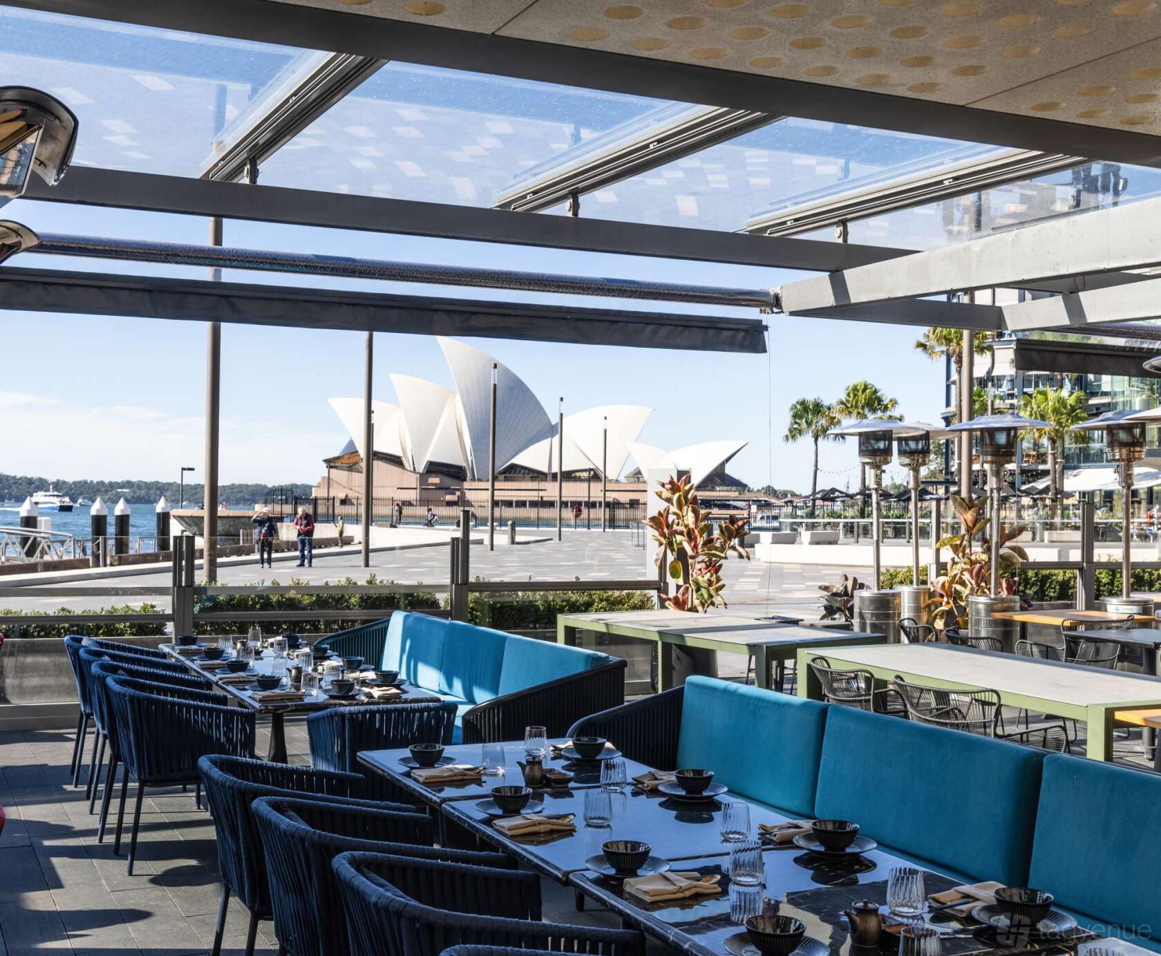 An open-air restaurant terrace with blue cushioned seating, set tables, and views of the Sydney Opera House at Luna Lu.