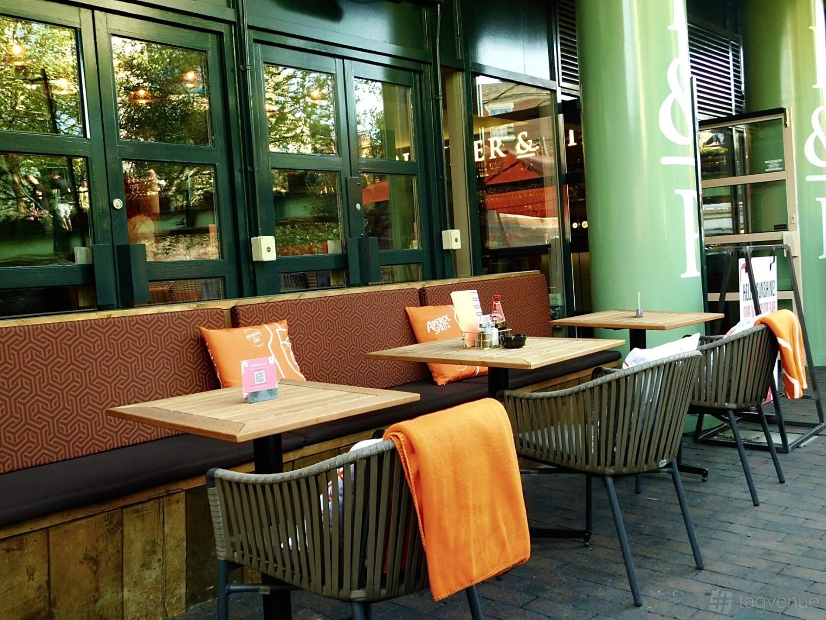 An outdoor bar area with wooden tables, cushioned bench seating, and orange blankets at Pitcher And Piano - Birmingham.