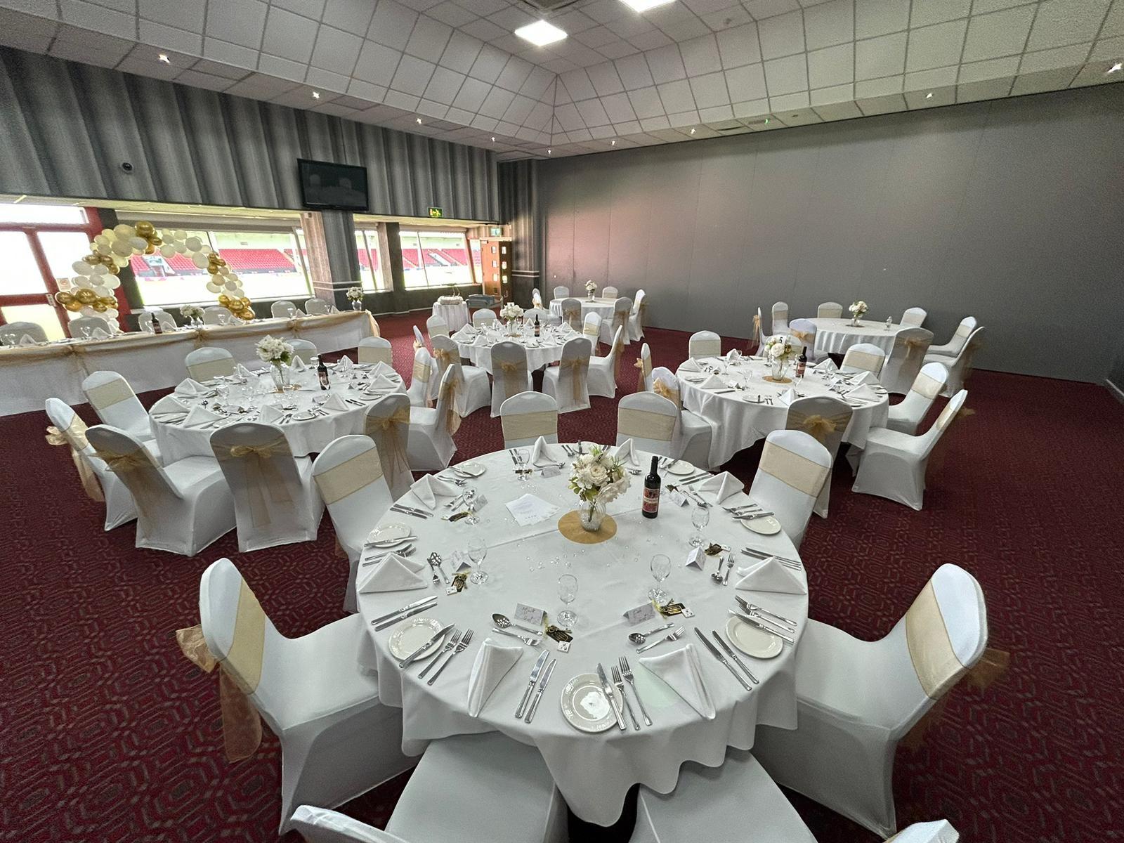 A lounge with round tables covered in white linens and gold chair sashes at Walsall Football Club.