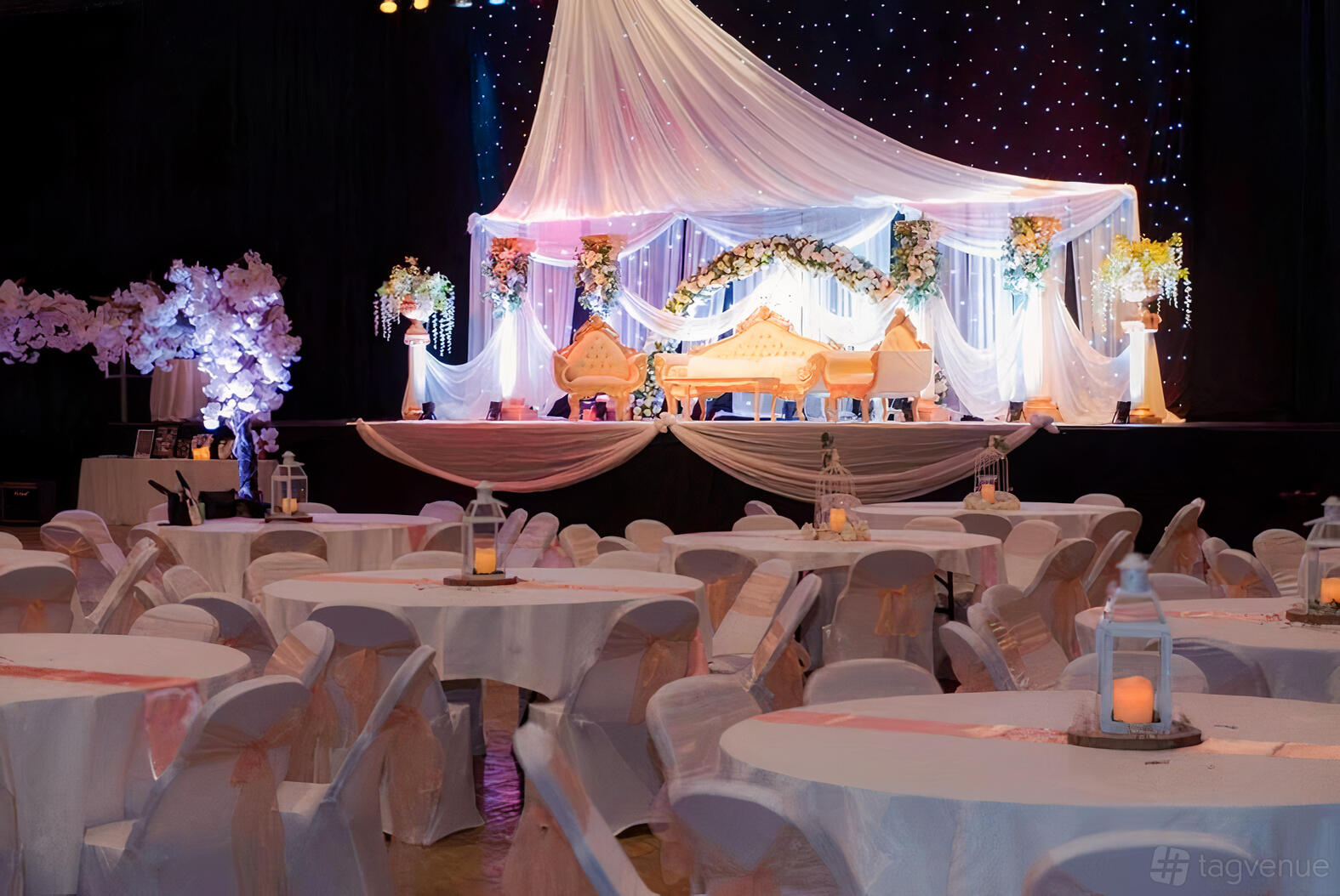 A museum hall with round tables draped in white linens and a decorated stage with floral arrangements at Colchester Weddings.