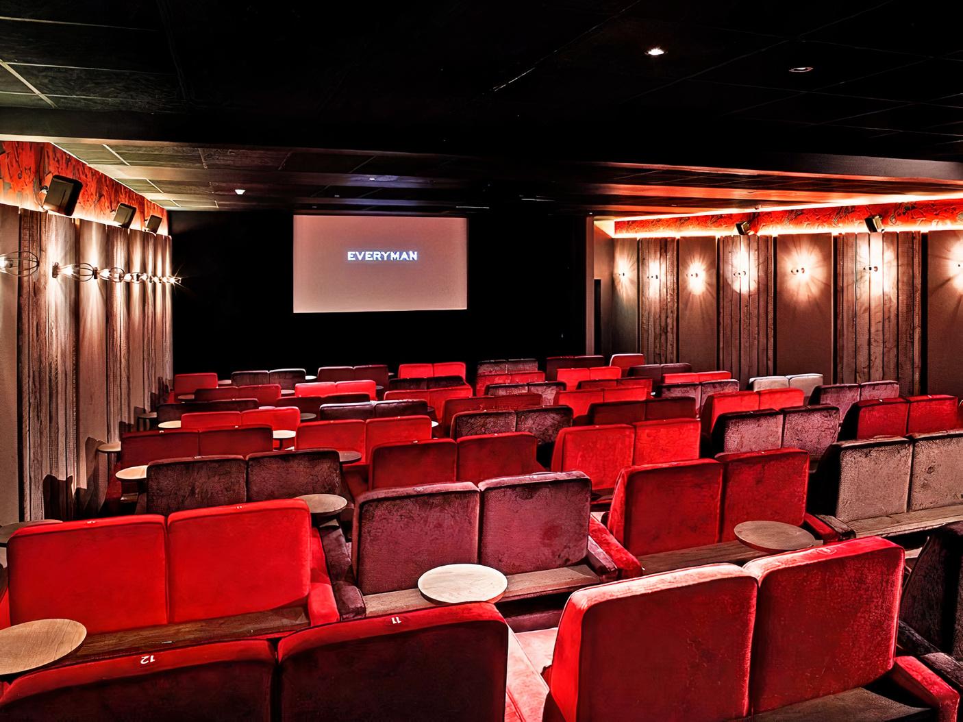 A screening room with red velvet armchairs, wooden tray tables, and wall sconces at Everyman Maida Vale.
