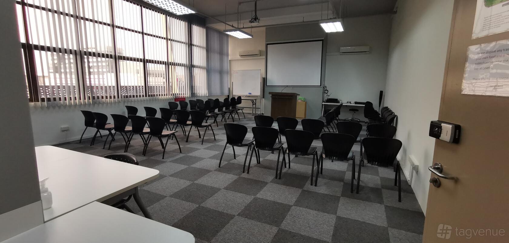 An auditorium with rows of black chairs, checkered carpet, a projector screen, podium, and large windows at Training Room at The Commerze.