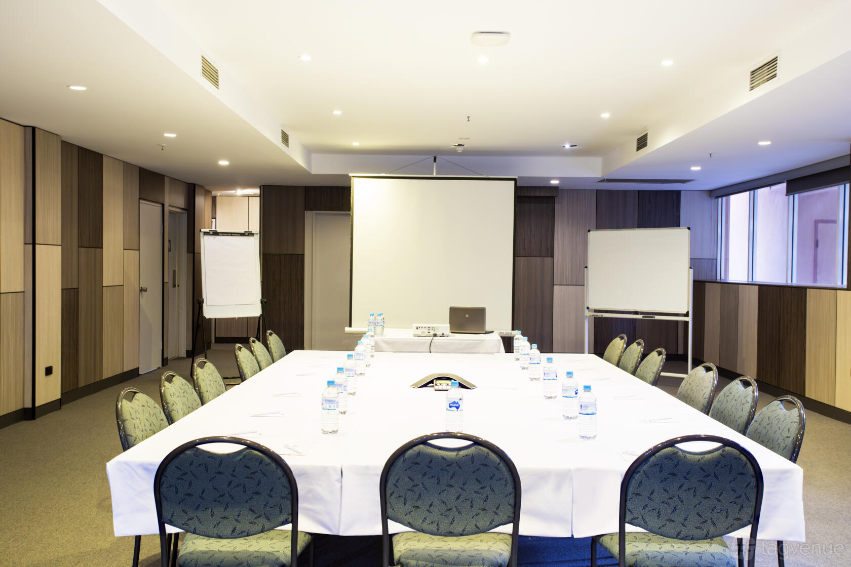A meeting room with a U-shaped table setup, projector screen, flip charts, and bottled water at Metro Aspire Hotel Sydney.