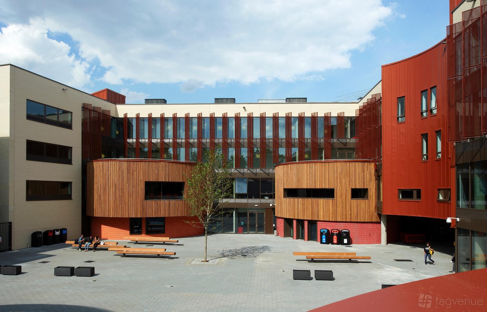 An academic venue with wood-paneled and red glass facades, central courtyard, and outdoor seating at ARU Venue Hire - Cambridge.