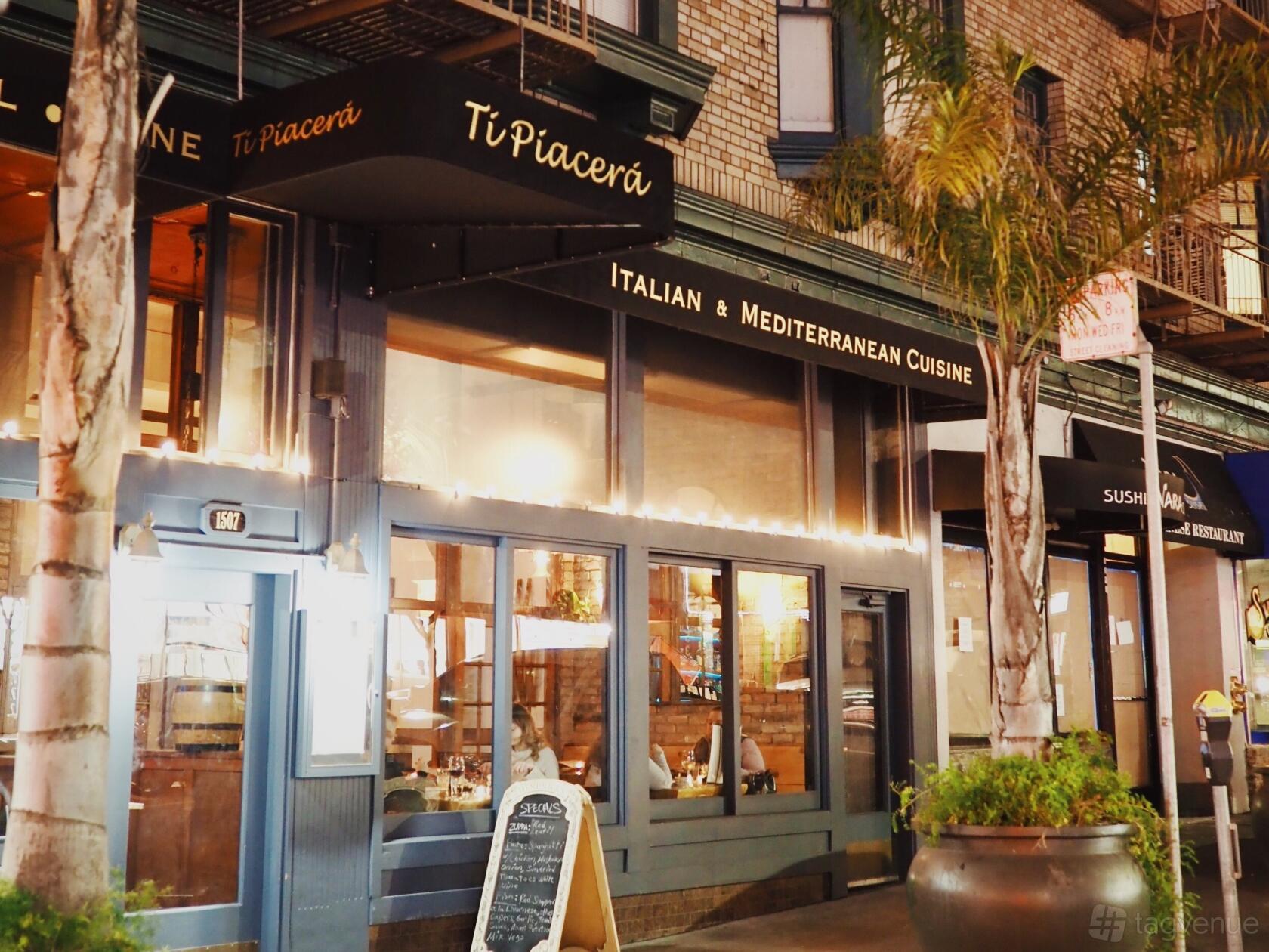 A restaurant exterior with large windows, illuminated signage, and palm trees at Ti Piacera.