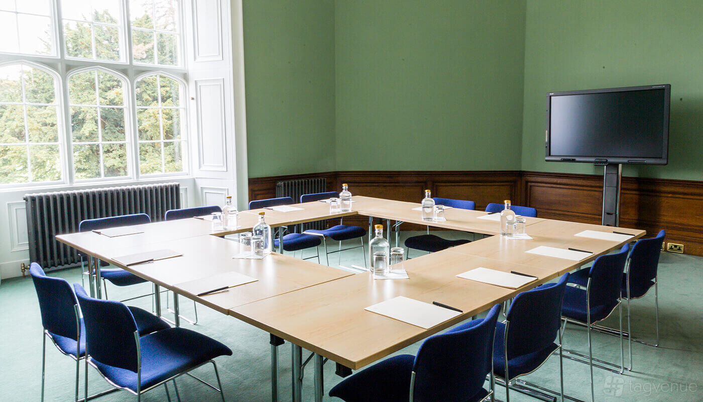 A castle meeting room with large windows, green walls, and U-shaped tables set with blue chairs at Ashridge House.