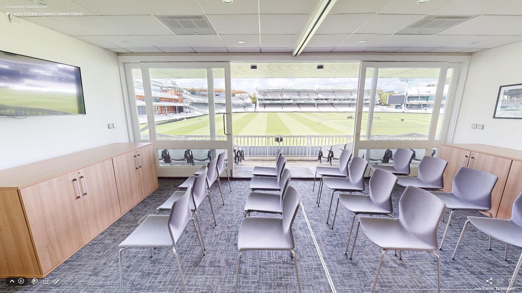 A meeting room with rows of gray chairs, large windows overlooking the cricket field at Lord's Cricket Ground.