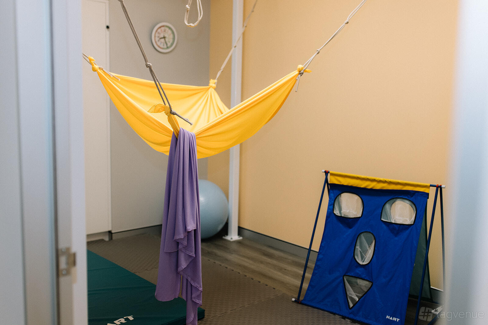 A therapy room with a yellow sensory hammock, padded flooring, and a blue sensory wall at Inside Out Therapy.