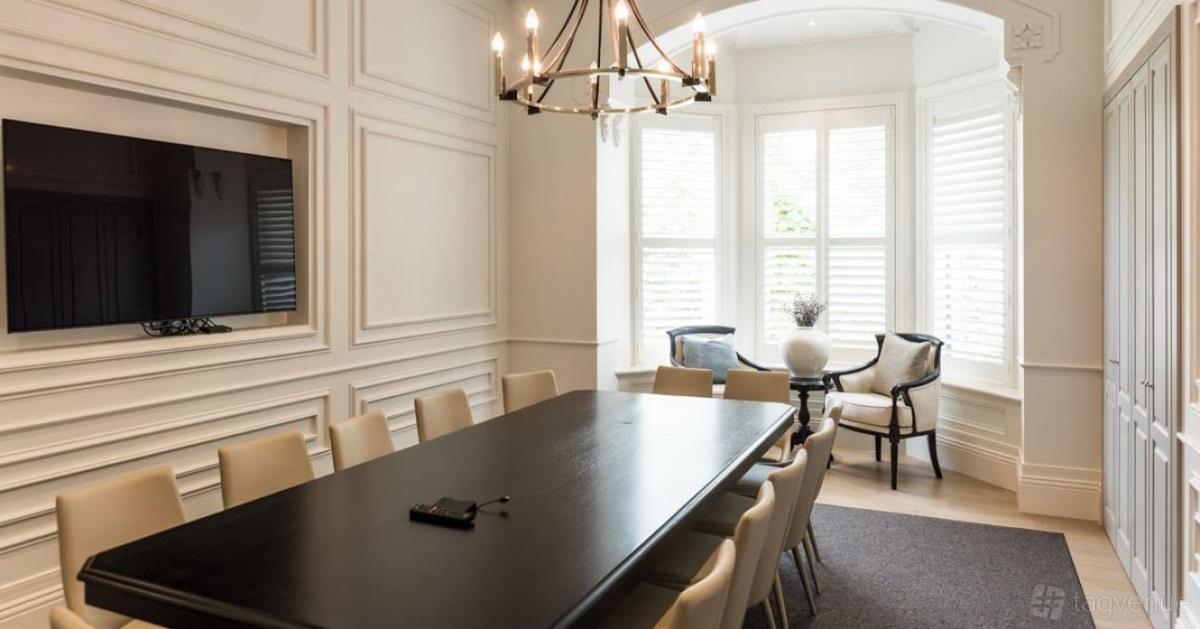 A dining room with a long black table, cream chairs, chandelier, and bay window seating at Marnong Estate.