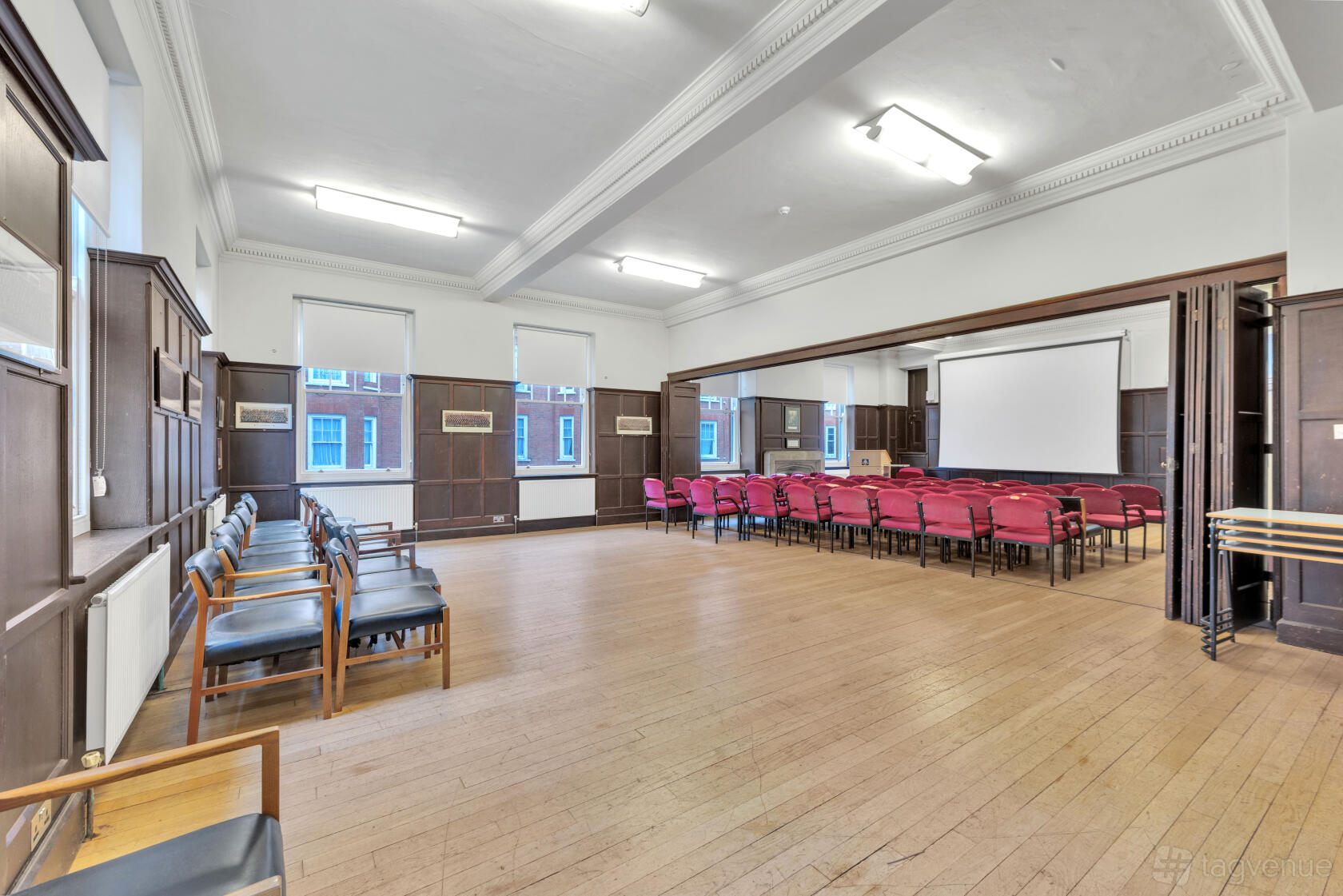 An auditorium with wood paneling, rows of red cushioned chairs, a projection screen, and natural light at Bloomsbury, Handel Street.