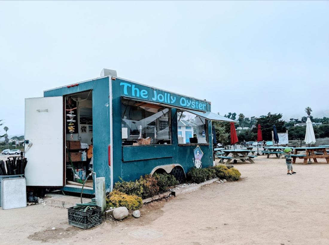 An outdoor bar with a blue food truck, picnic tables, and surrounding umbrellas at The Jolly Oyster Shuck Shack.
