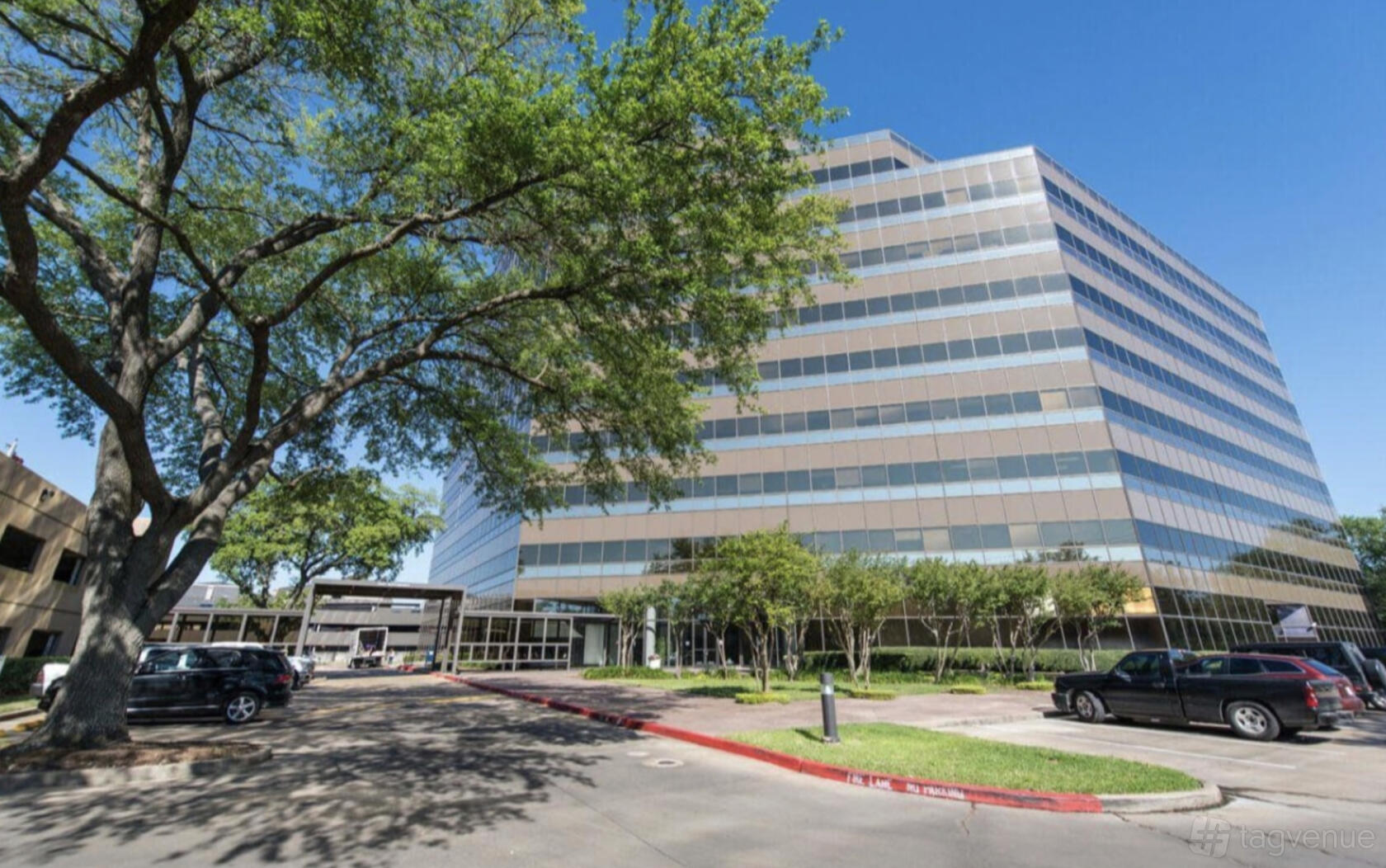 An office building with large glass windows, mature trees, and a parking lot at Private Office Westchase.