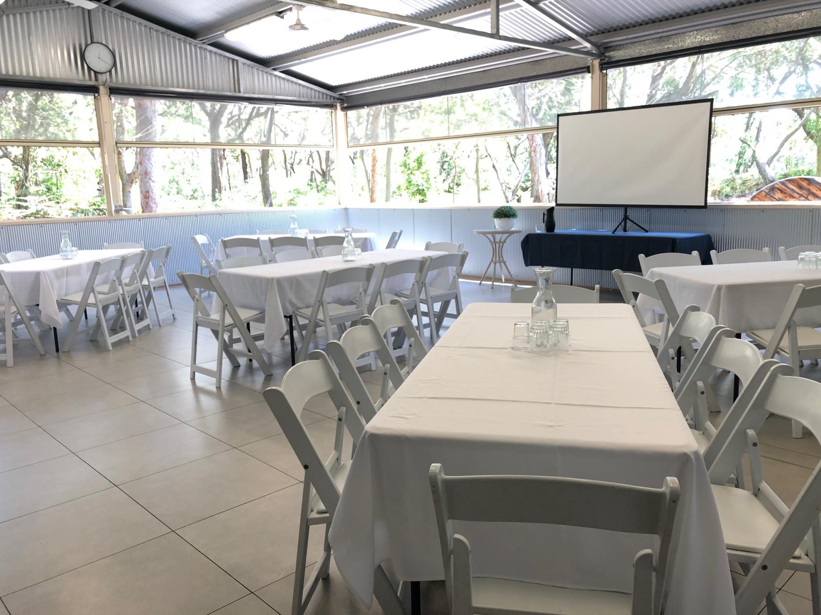An event room with white folding chairs, round and rectangular tables with white linens, and a projector screen at The Retreat Port Stephens.