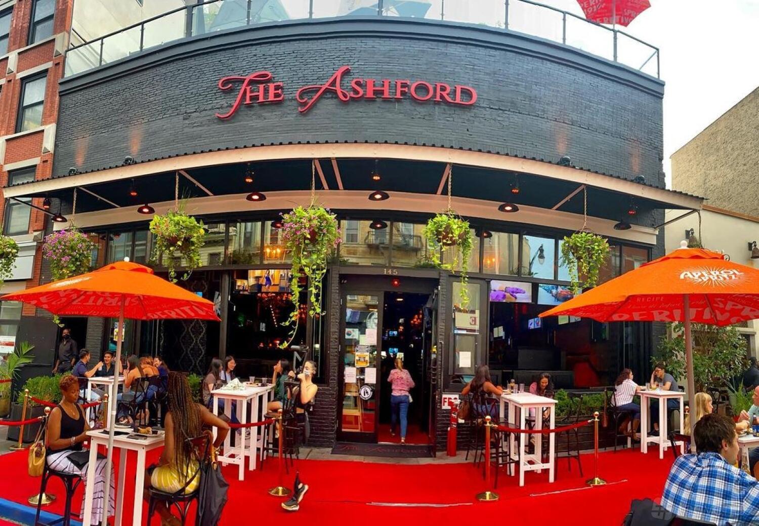 An outdoor terrace with red umbrellas, hanging plants, and high tables in front of The Ashford.