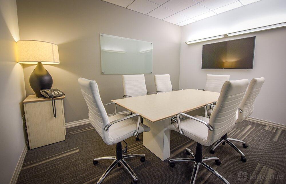 A meeting room with a rectangular table, six white chairs, a wall-mounted TV, and a glass board at Premier Workspaces - Koll Center.