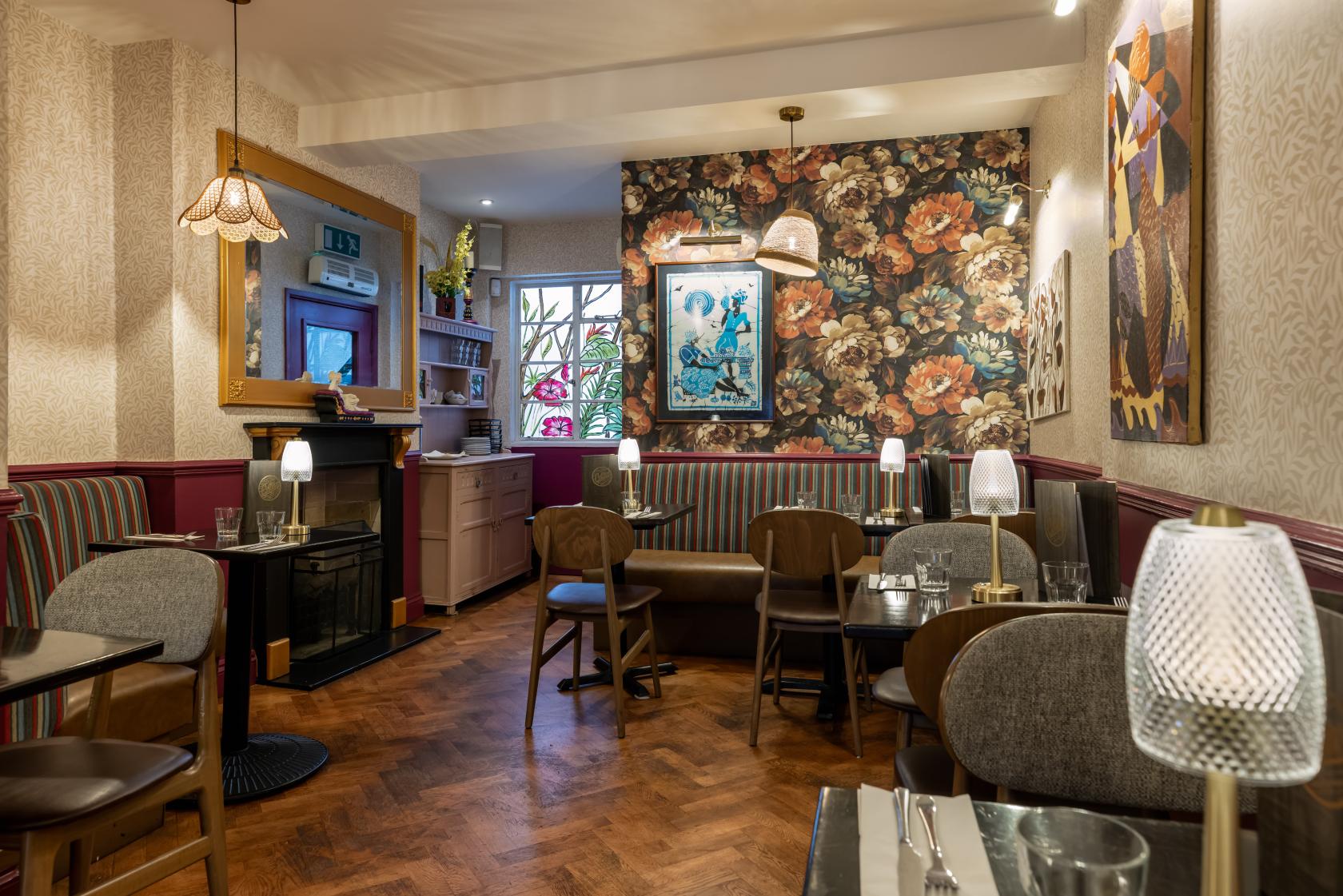 A dining room with patterned wallpaper, striped banquette seating, and round tables with glass lamps at Cottons Camden.