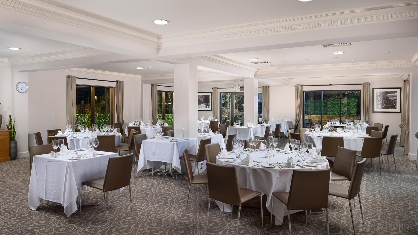 An event space with round tables draped in white linens, brown chairs, and large windows at Voco Oxford Spires.