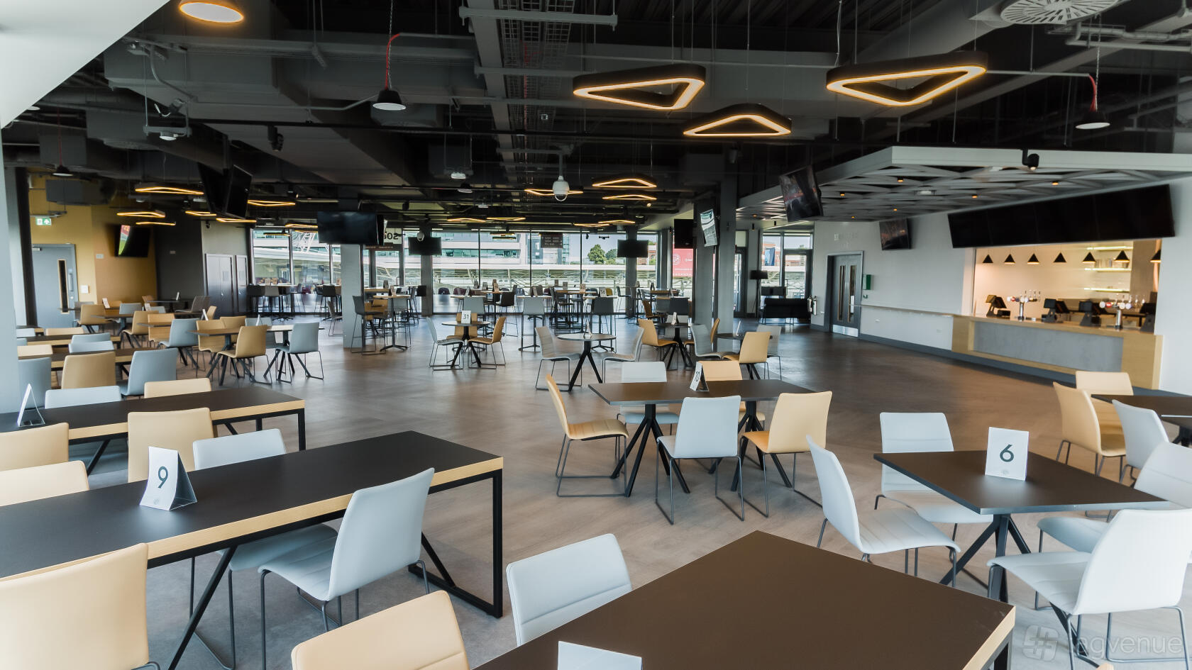 A spacious lounge with geometric ceiling lights, floor-to-ceiling windows, and tables with mixed chairs at Brentford Football Club.