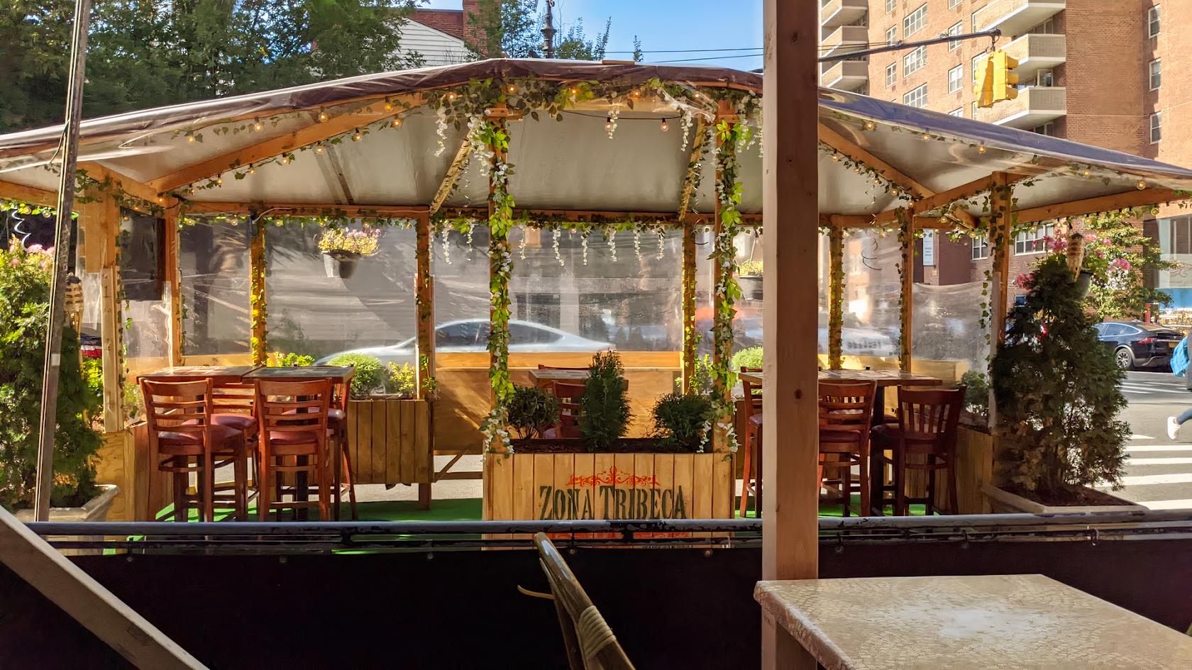An outdoor restaurant patio with wooden tables, chairs, hanging greenery, and clear side panels at Zona Tribeca.