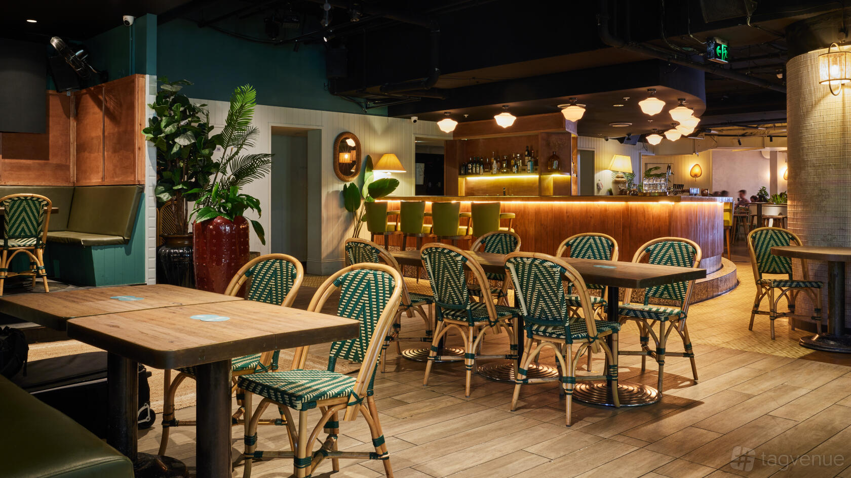 A pub dining room with wooden tables, green wicker chairs, potted plants, and a curved bar at Kent St. Hotel.