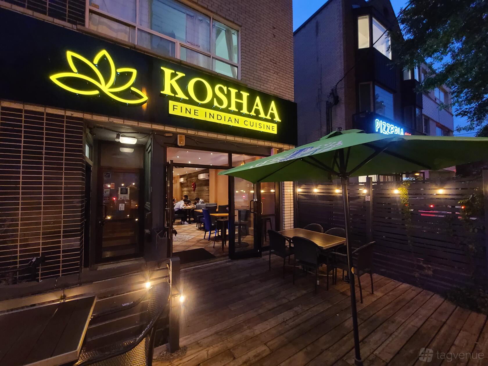 A restaurant with an outdoor wooden deck, umbrella-shaded tables, and illuminated signage at Koshaa Fine Indian Cuisine.
