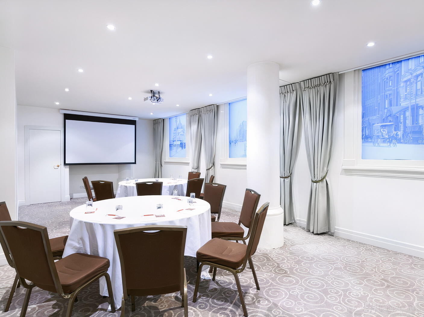 A meeting room with round tables covered in white linens, cushioned chairs, and a projector screen at Rendezvous Hotel Melbourne.