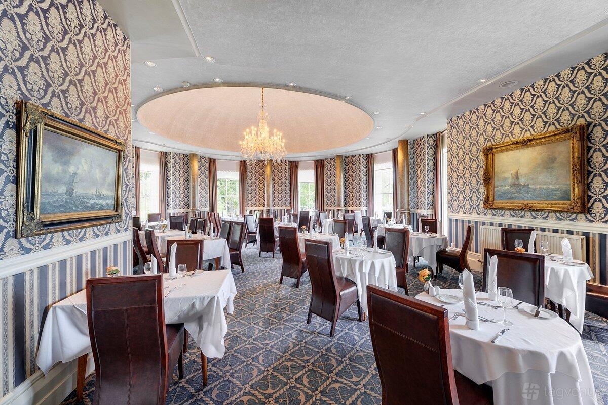 A dining room with patterned wallpaper, crystal chandelier, round tables with white linens, and large windows at The Duke of Cornwall Hotel.