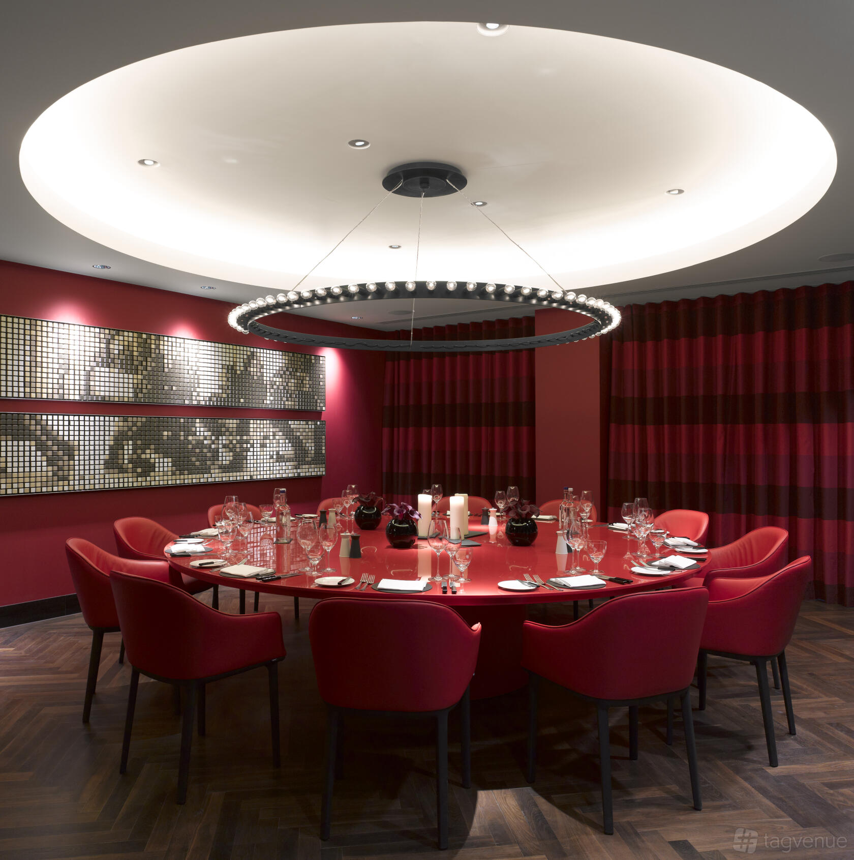 A dining room with a large oval red table, red chairs, and a circular chandelier at South Place Hotel