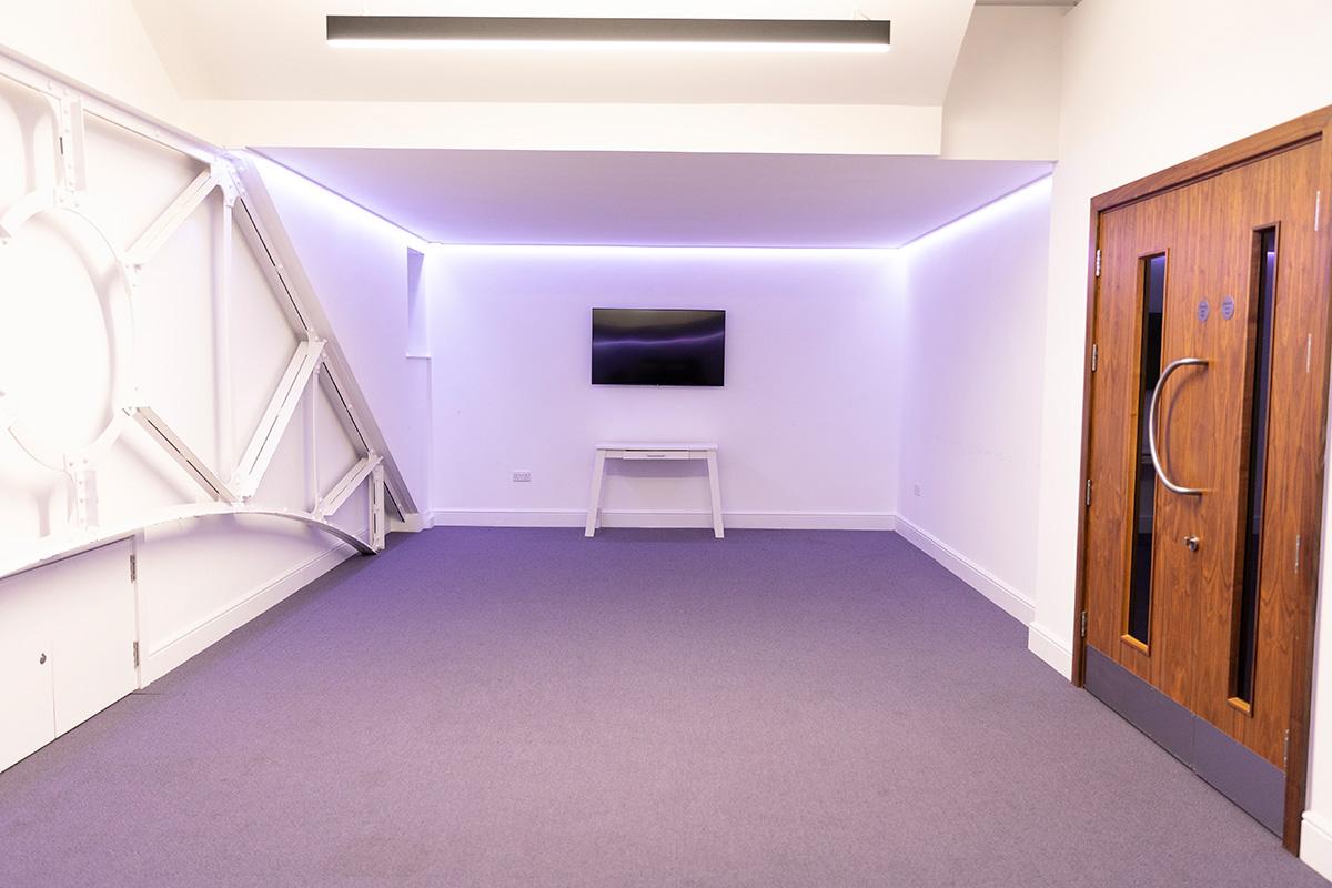 A meeting room with a wall-mounted TV, white accent lighting, and carpeted floor at The Tab Centre.