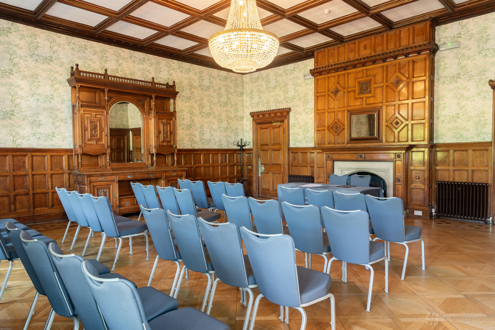 An event space with wood paneling, a large chandelier, patterned wallpaper, and rows of blue chairs at Pendley Manor Hotel.