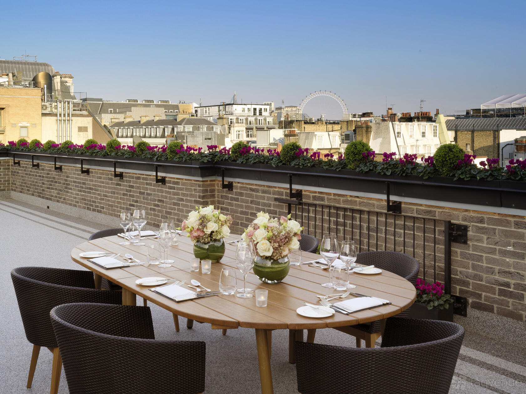 A hotel rooftop terrace with an oval table set for dining, wicker chairs, and flower arrangements at The Stafford London.