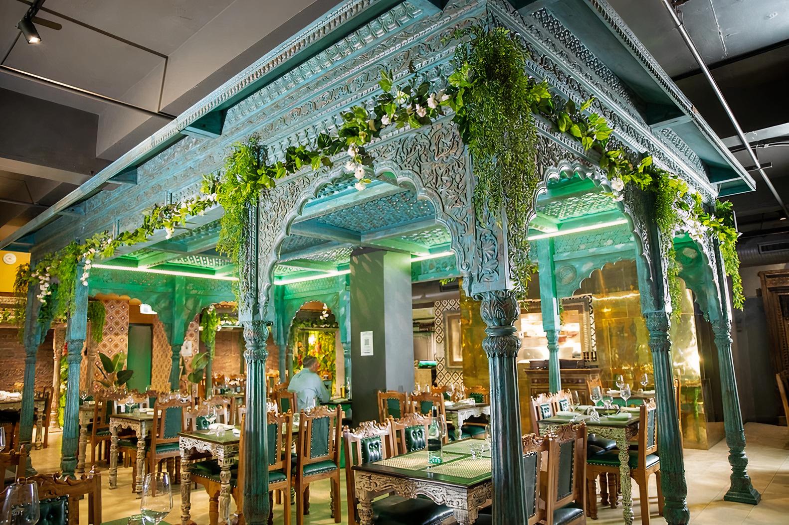 A restaurant with ornate carved turquoise arches, hanging greenery, and wooden tables at The Grand Palace - Indian Restaurant (Sydney).