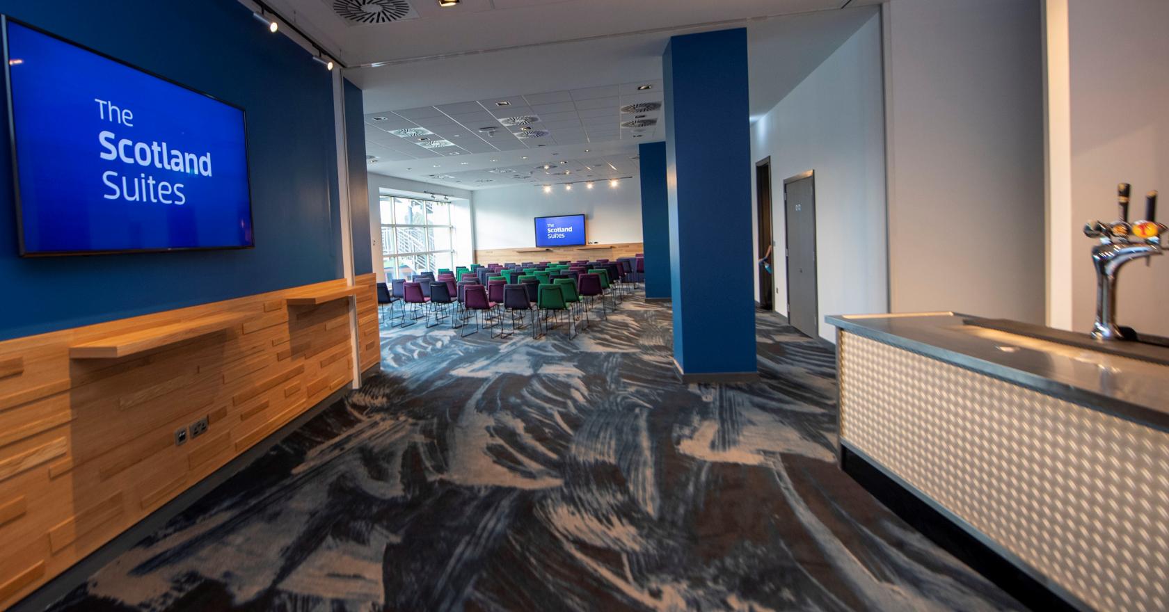 A meeting room with rows of colorful chairs facing a presentation screen and large windows at BT Murrayfield Stadium.