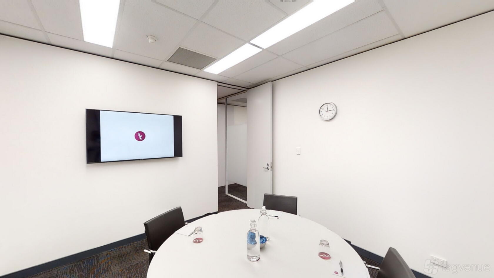 A meeting room with a round table, black chairs, wall-mounted screen, and clock at Karstens Perth.