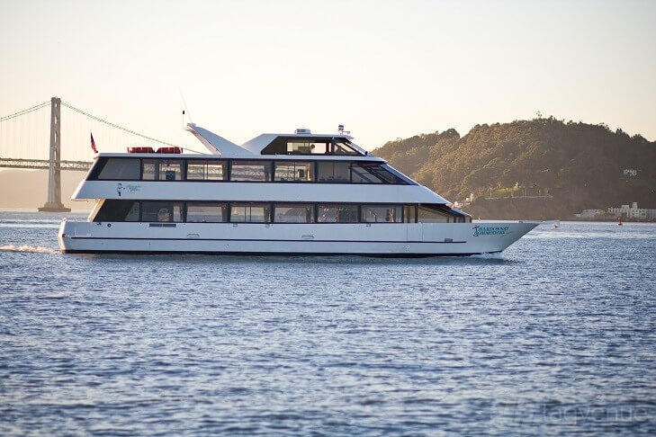 An event venue boat with three decks and large windows cruising on the water near a bridge at Chardonnay.