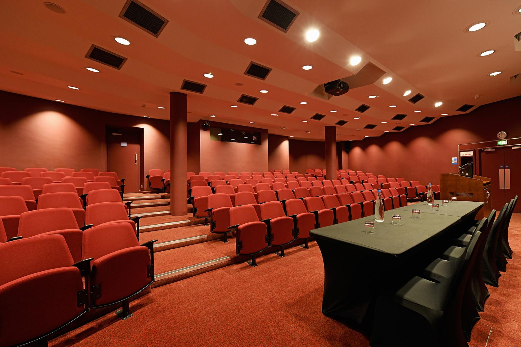 An auditorium with tiered red seating, wall-mounted lights, and a panel table with microphones at SCI Belgravia.