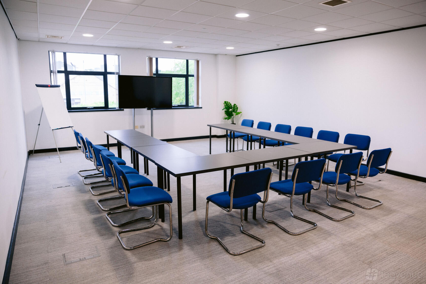 A meeting room with U-shaped tables, blue chairs, large windows, a TV screen, and a flipchart at Lawrence House - Hub26.