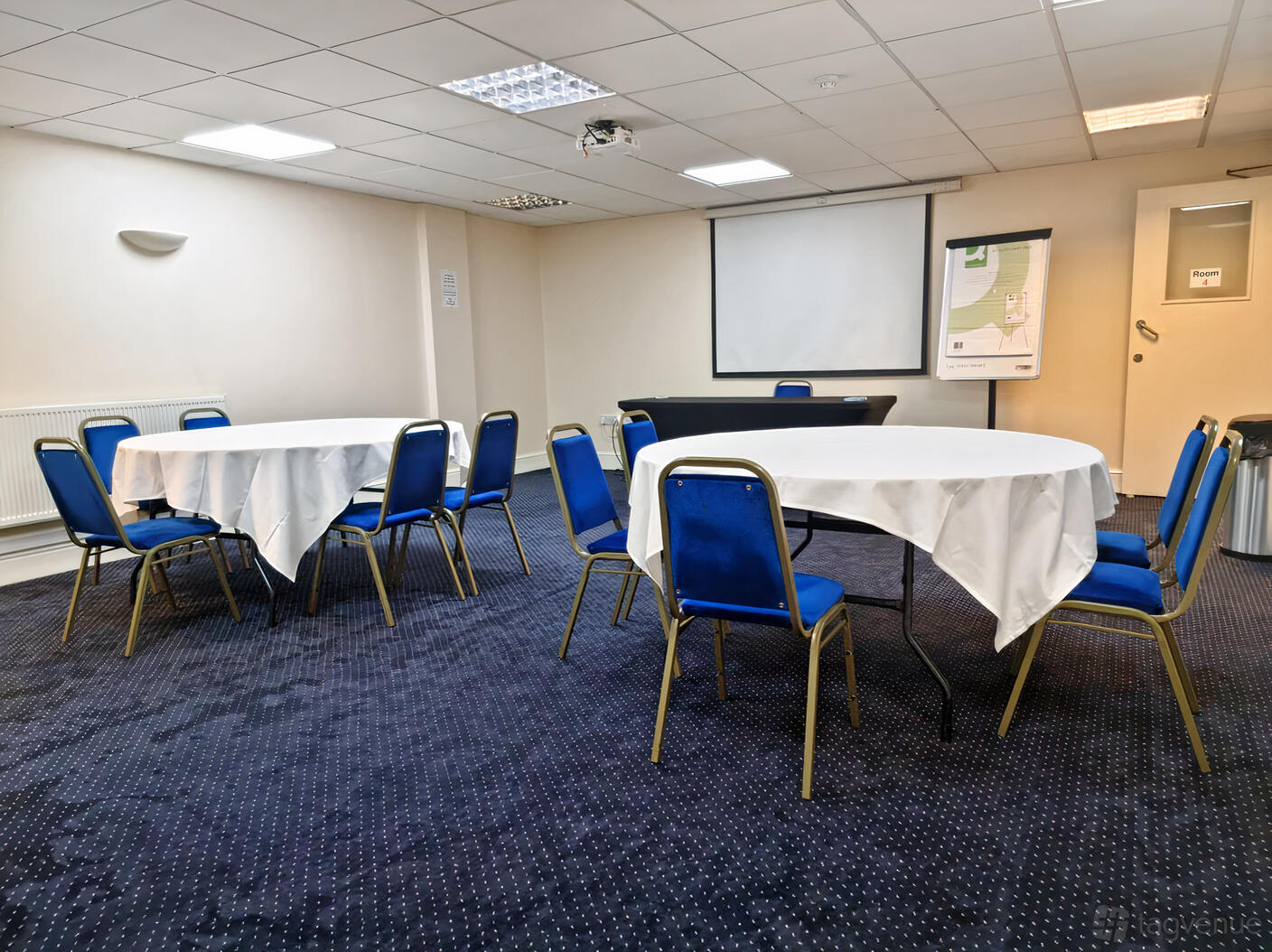 A meeting room with round tables covered in white linens, blue chairs, and a projector screen at The Saffron Centre.