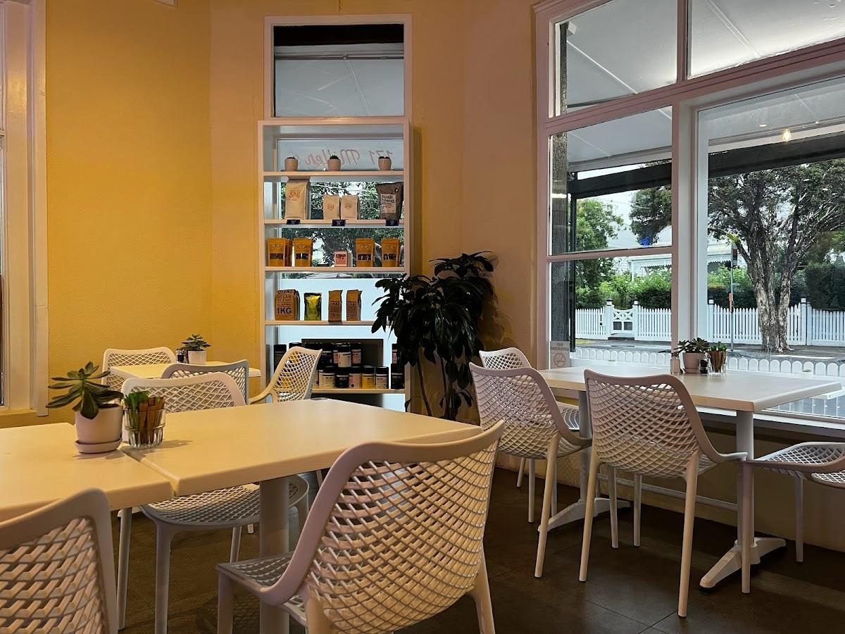 A cafe with white mesh chairs, potted plants on tables, large windows, and a display shelf at Twice Shy.