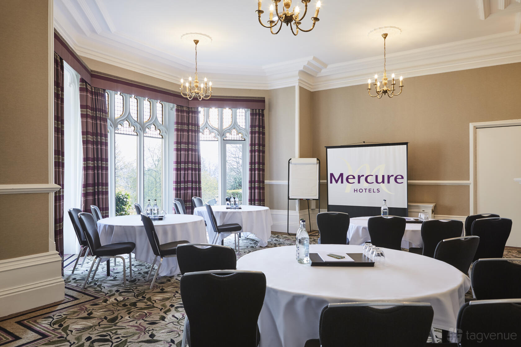 A function room with round tables, black chairs, chandeliers, and bay windows at Mercure Sheffield Kenwood Hall Hotel & Spa.