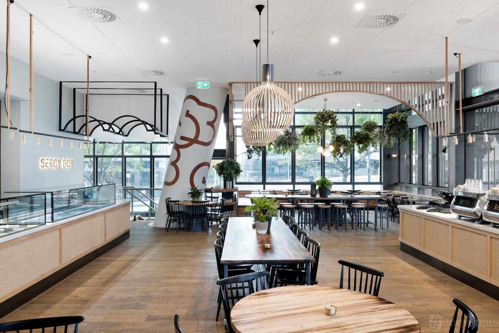 An event space with large windows, wooden floors, pendant lights, and greenery at Sergy Boy.