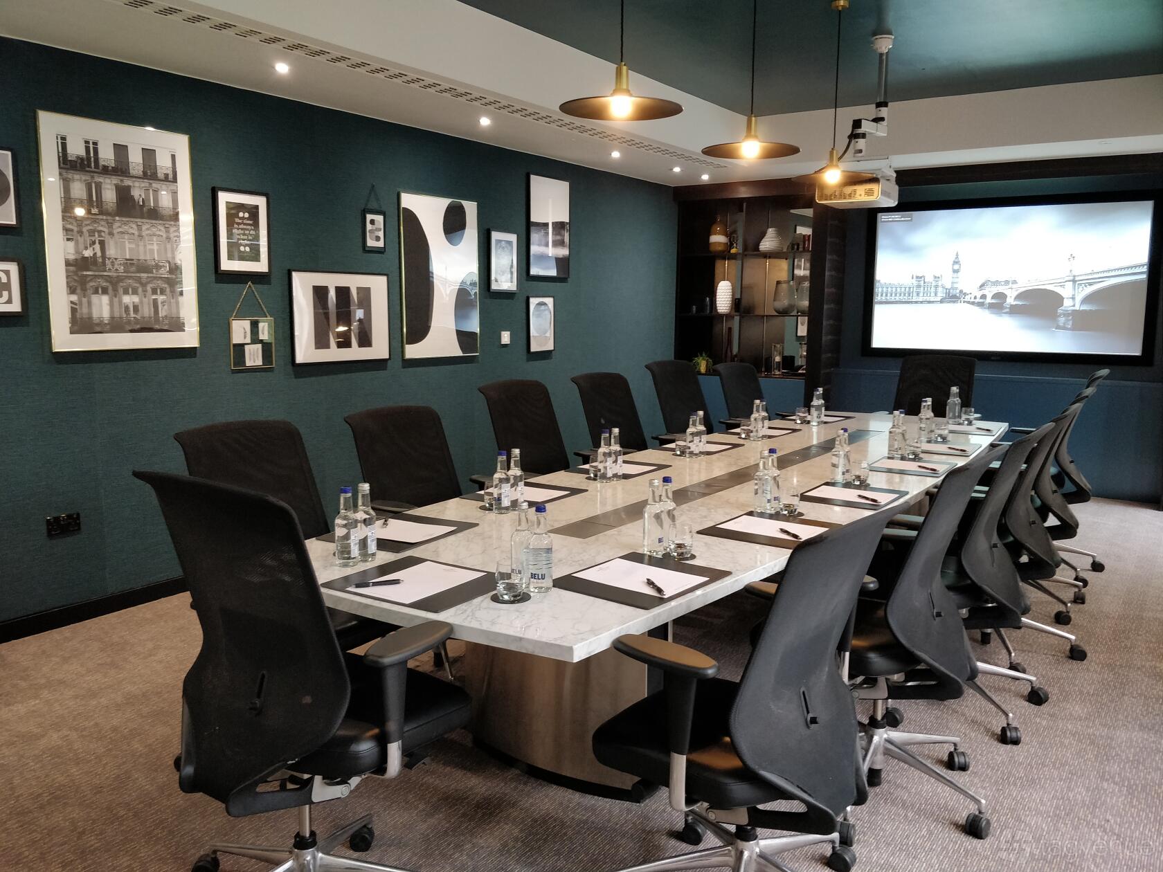 A hotel boardroom with a marble conference table, ergonomic chairs, wall art, and a projector screen at DoubleTree by Hilton London Kensington.