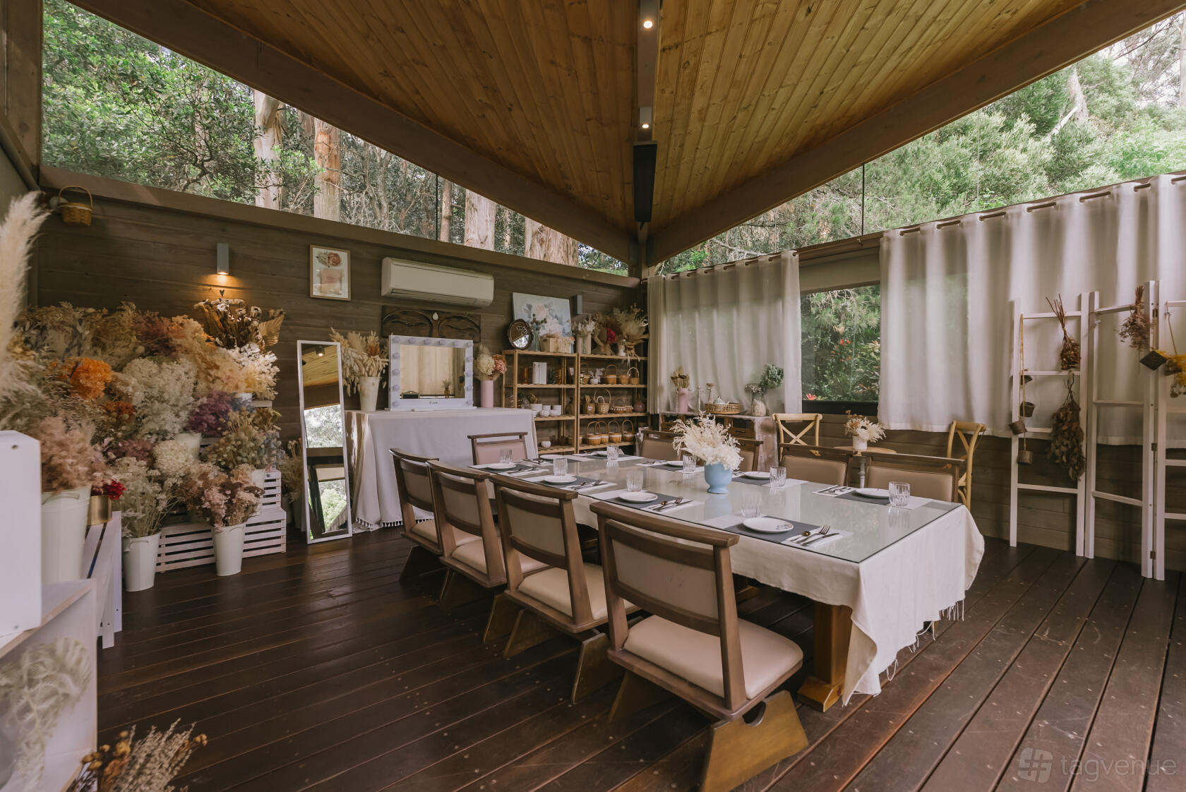 A restaurant dining room with a long glass table, wooden ceiling, dried floral arrangements, and large windows at Olinda Tea House.