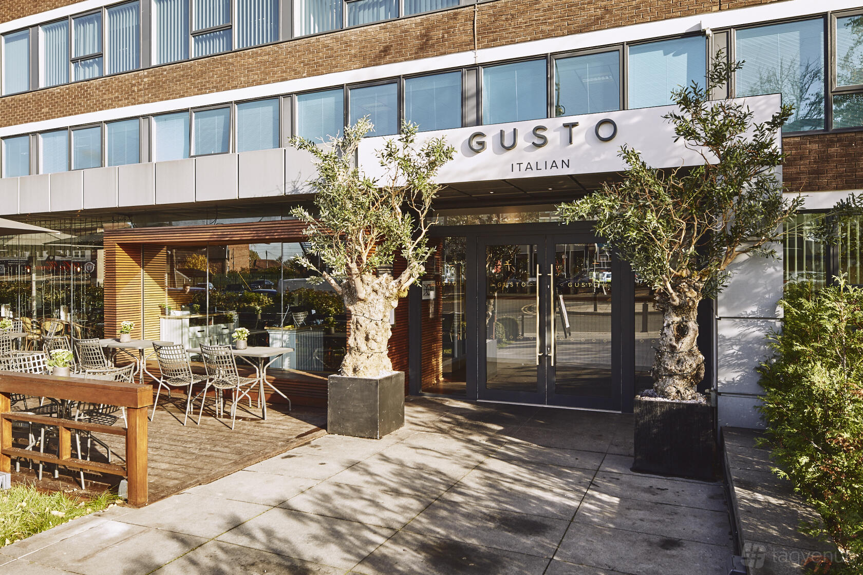 A restaurant entrance with large glass doors, potted olive trees, and outdoor patio seating at Gusto Italian Cheadle.