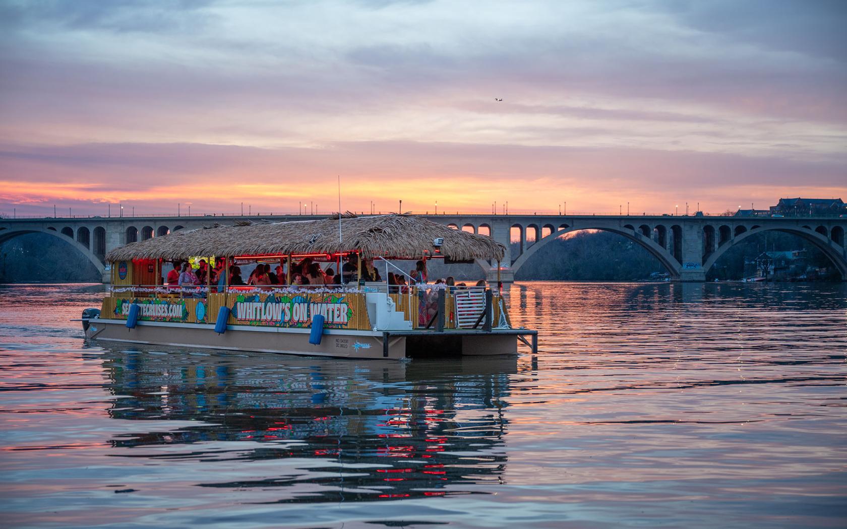 A boat with a thatched roof and open-air seating cruising at sunset near a bridge at Whitlow's on the Water.