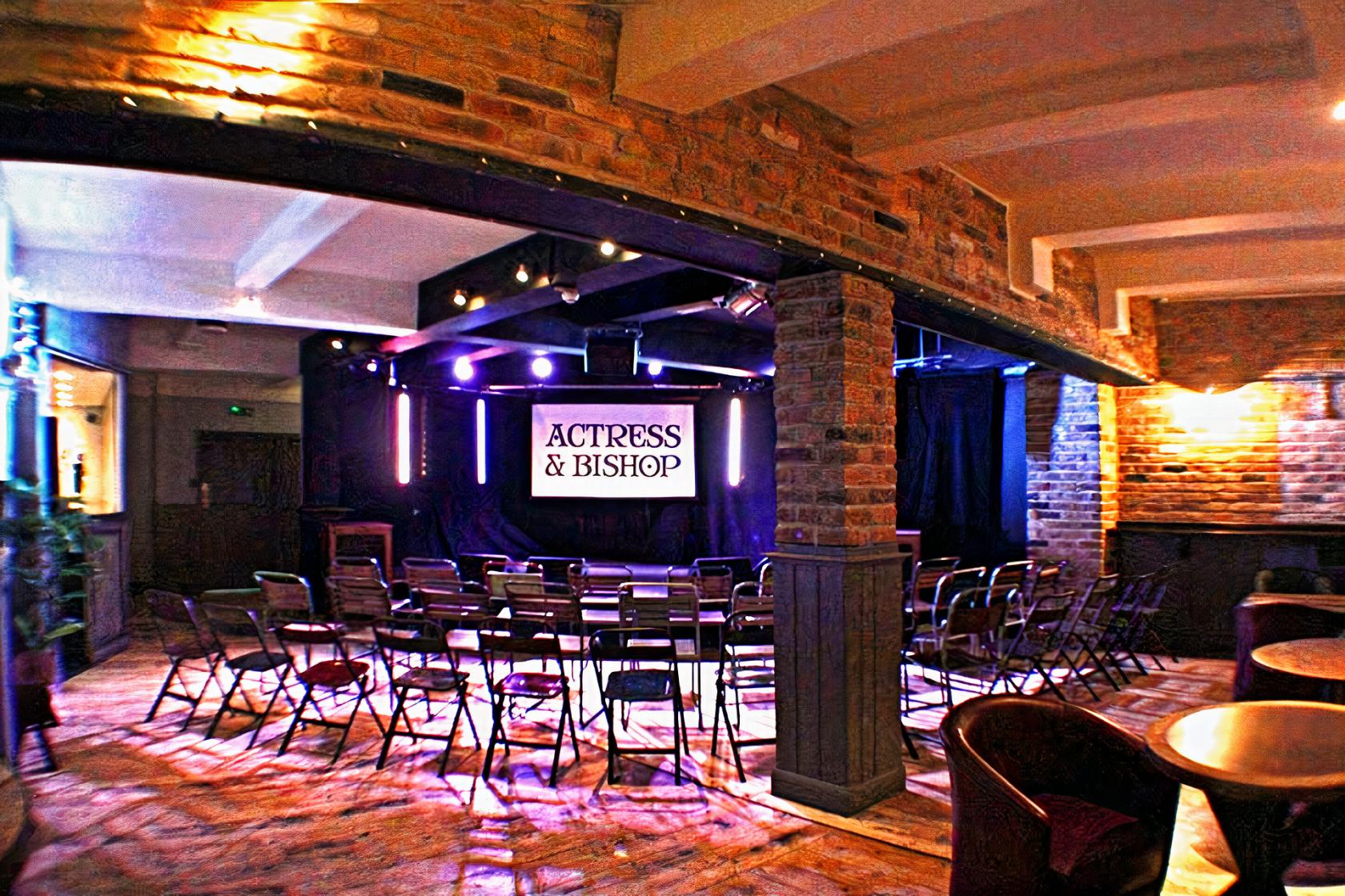 An upstairs event space with exposed brick arches, stage lighting, and rows of folding chairs at The Actress & Bishop.