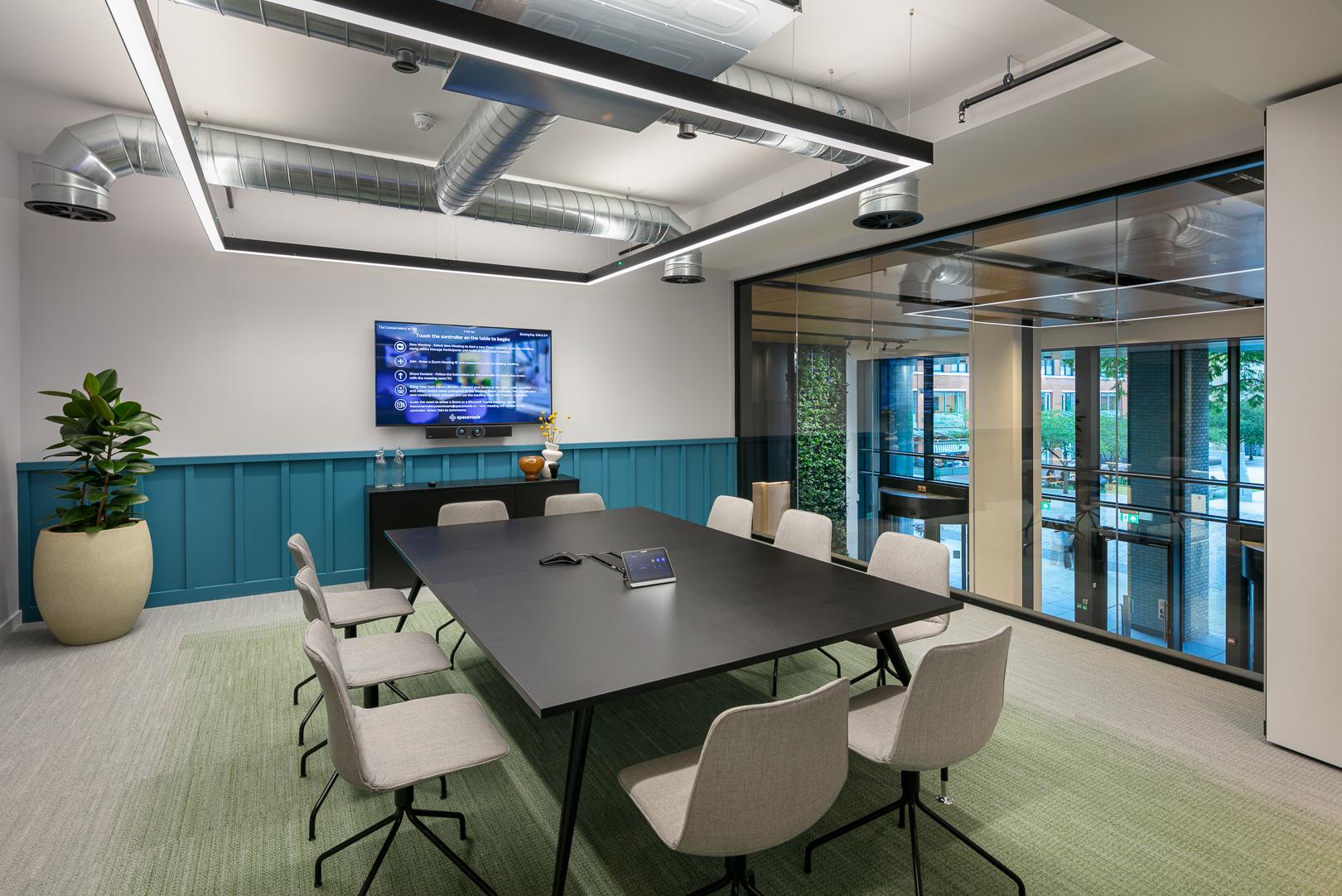 A meeting room with a black conference table, beige chairs, glass wall, and potted plant at 10X by Spacemade.